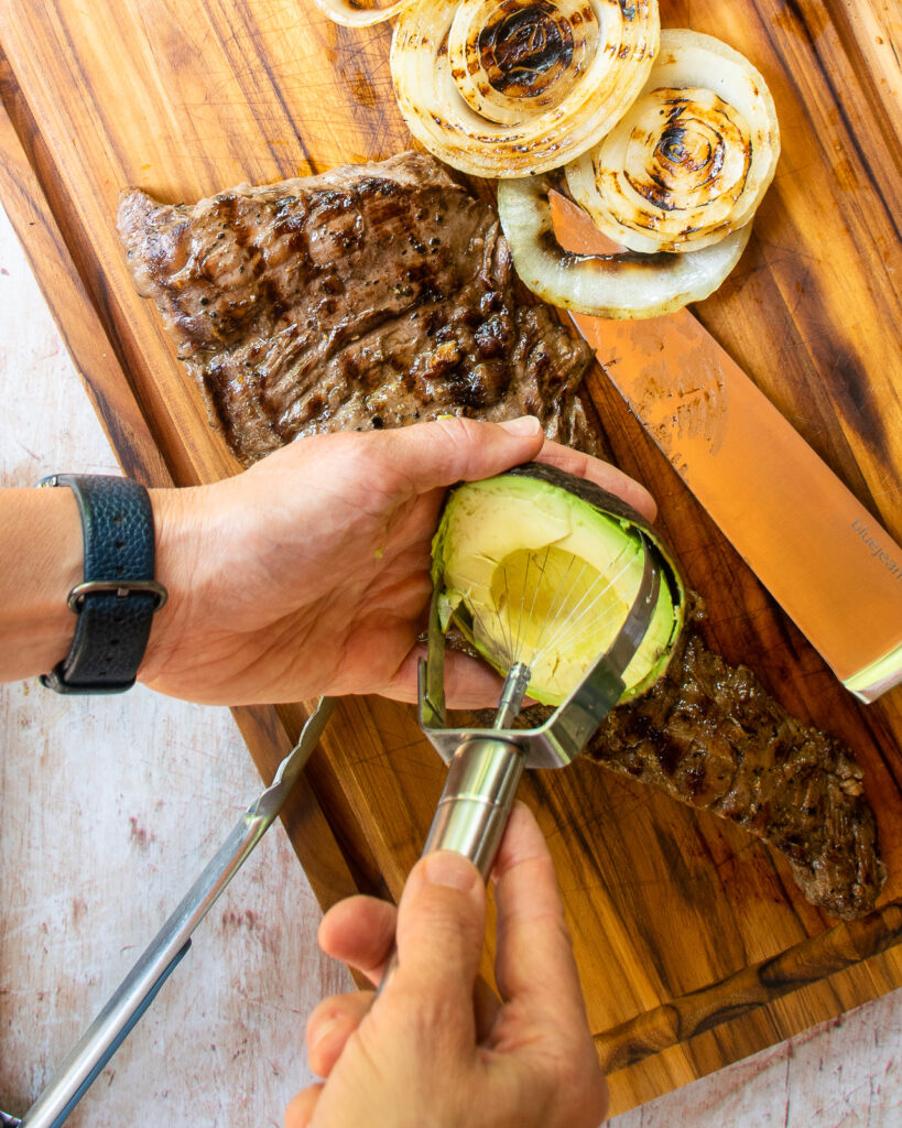 Hands using an avocado slicer to slice avocado with grilled skirt steak and grilled onions on a cutting board near by.