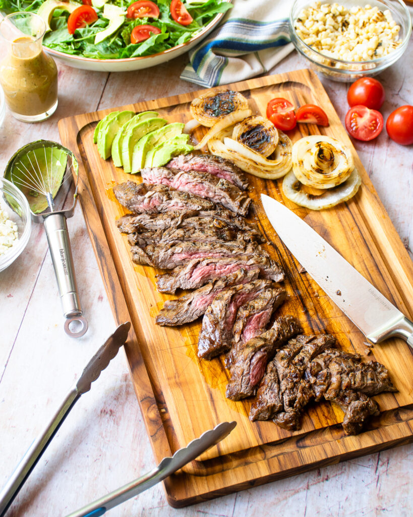 Looking down on ingredients for a grilled steak salad - grilled skirt steak on a cutting board, onions, tomatoes, avocado, grilled corn, blue cheese crumbles and a bowl of greens.