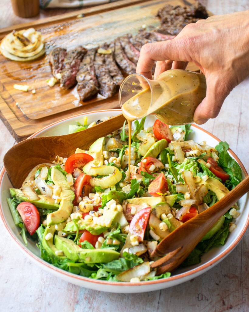 A hand pouring creamy balsamic dressing over a bowl of salad with a grilled skirt steak in the background on a cutting board.