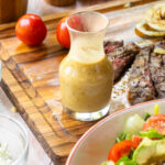 A close up of creamy balsamic dressing on a cutting board with grilled steak in the background and a bowl of salad in the foreground.