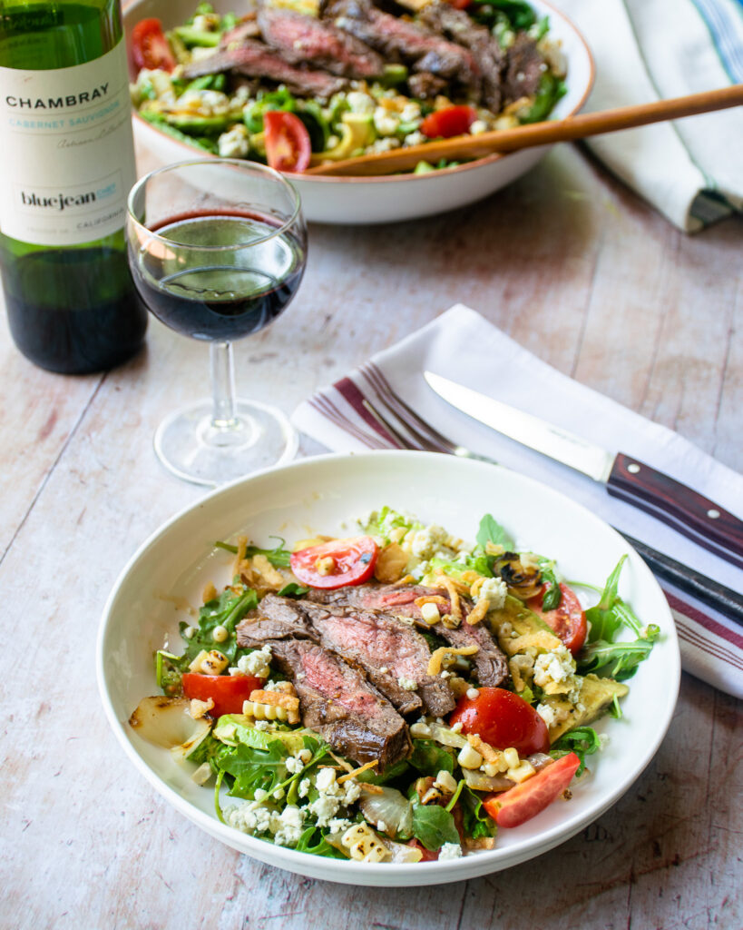 A white bowl with grilled steak salad, a place setting and a glass and bottle of wine.