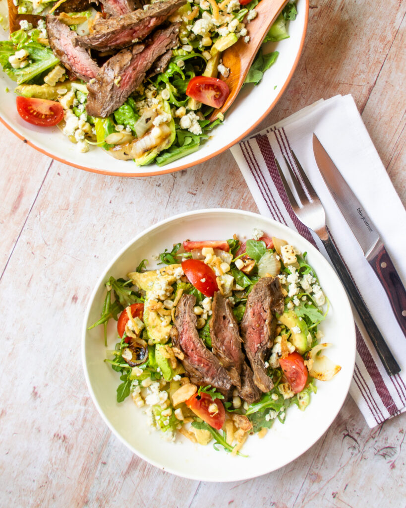 Looking down on a white bowl with grilled steak salad, a place setting and a larger serving bowl with more salad inside.