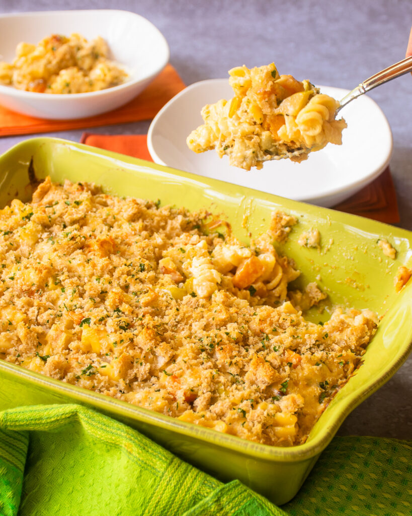 A spoon lifting a portion of butternut squash apple mac and cheese above a green casserole with white plates in the background.