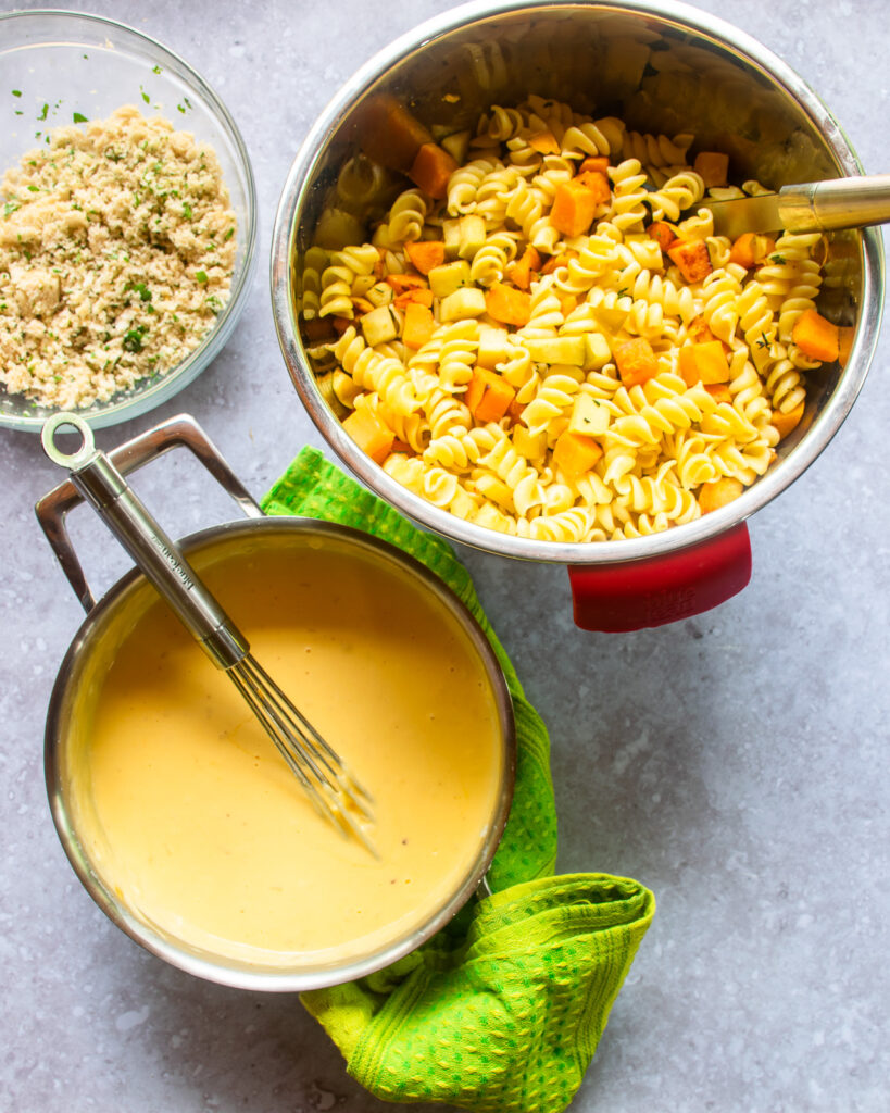 Looking down on a saucepan with cheese sauce and a whisk next to a bowl with pasta, butternut squash and apple inside and a second bowl with crumb topping.
