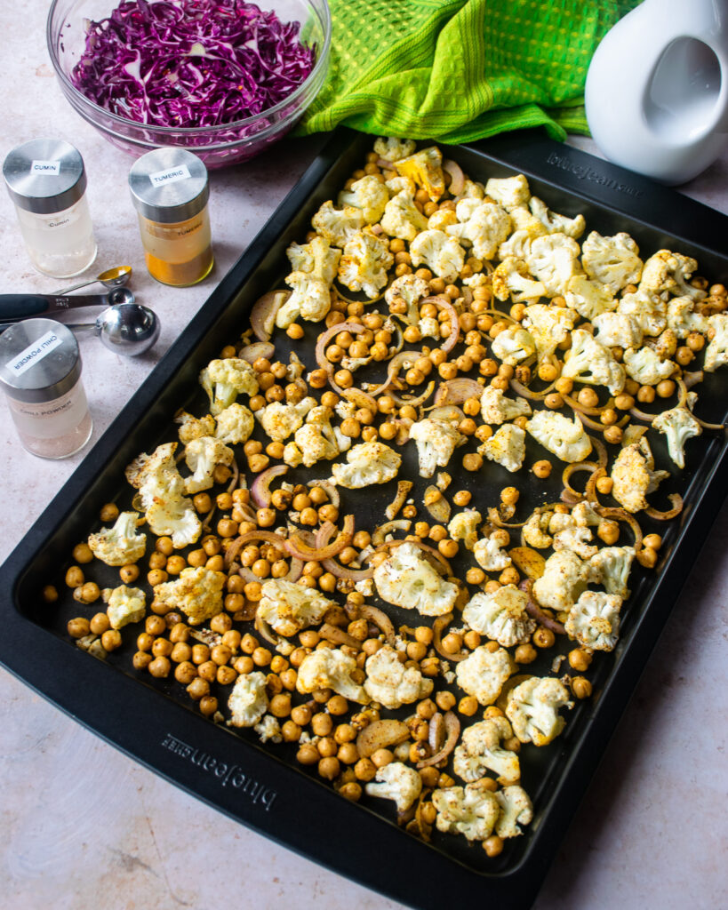 raw cauliflower and chickpeas tossed with spices on a baking sheet with a bowl of pickled red cabbage and spice jars near by.