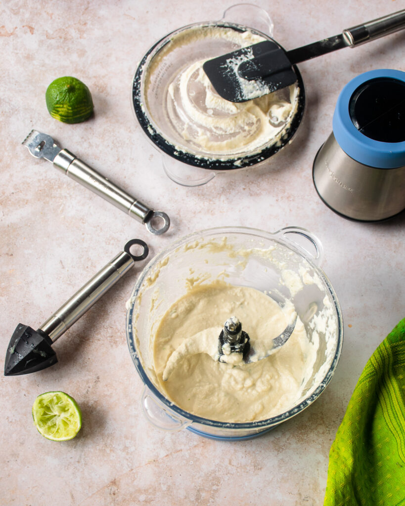 Looking down into a food processor with cashew crema inside, a reamer, a zester and some spent lime halves near by.