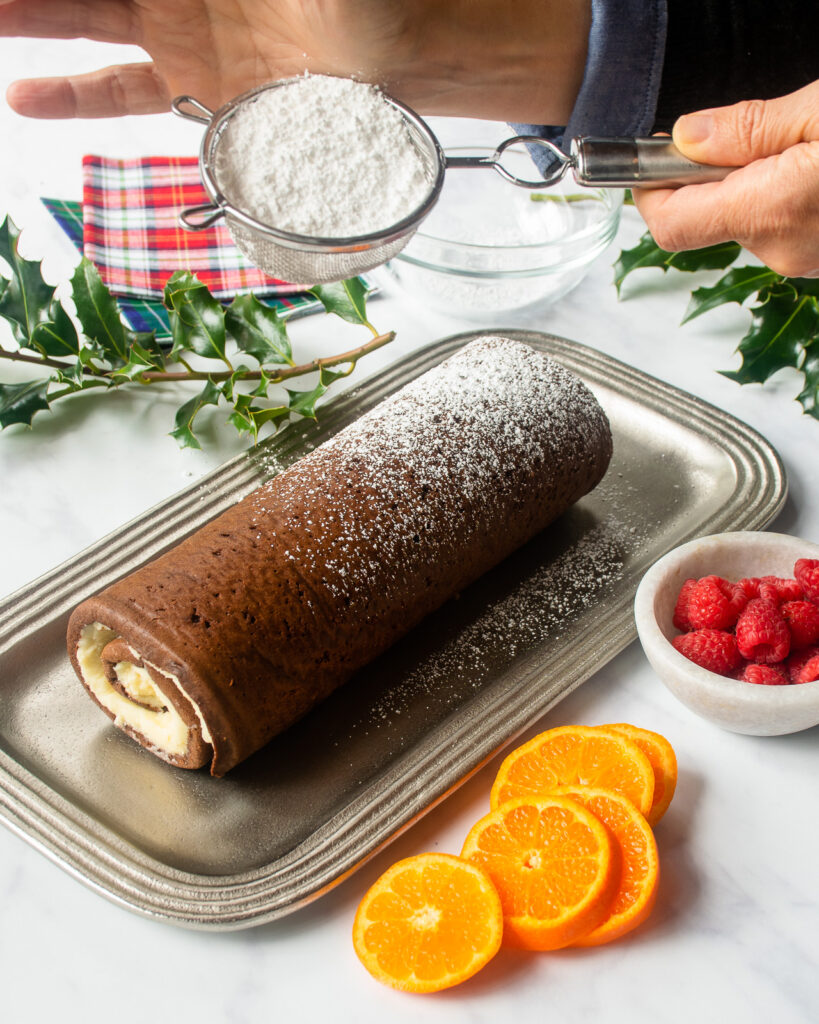 A hand dusting a chocolate swiss roll with powdered sugar using a small strainer.