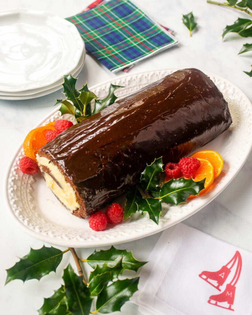 a chocolate coated Chocolate Swiss Roll on a white plate with mandarin orange slices, raspberries and holly decorating it.