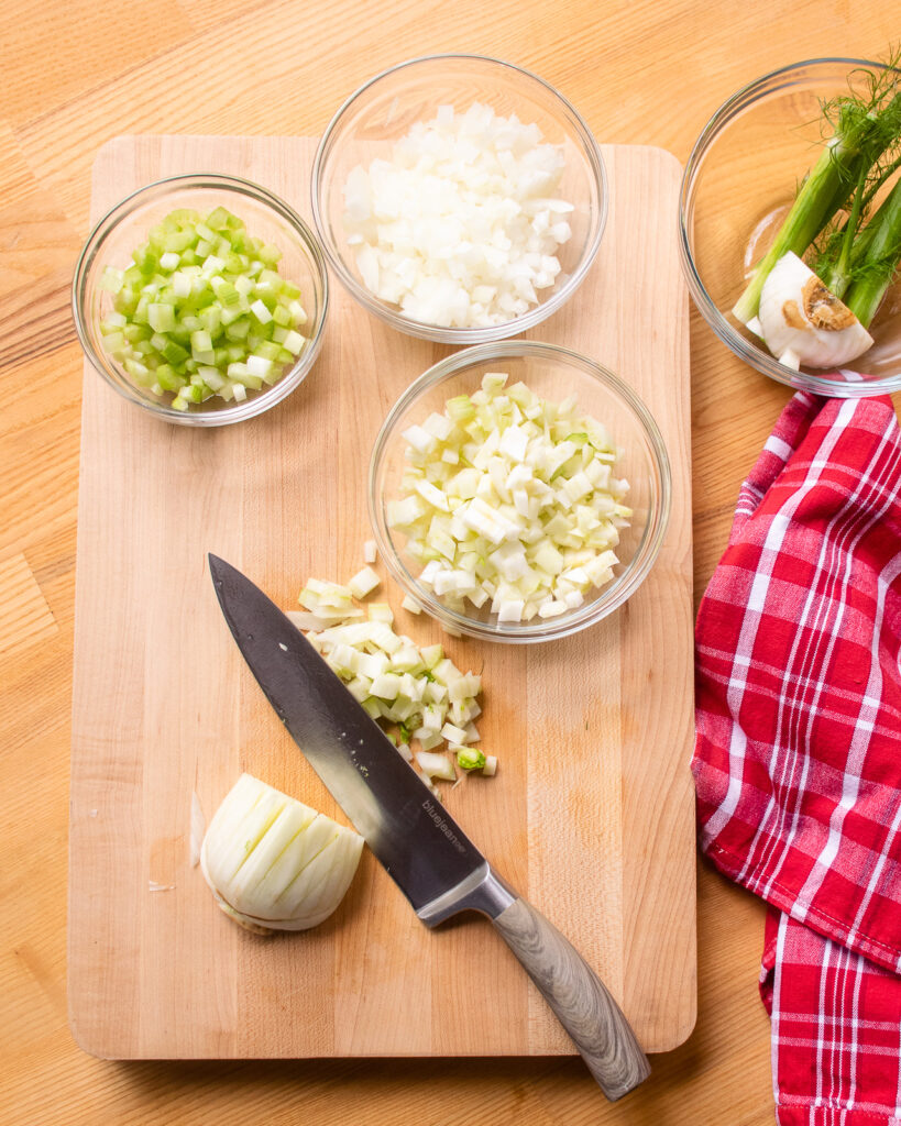 A cutting board on a wooden countertop with a bowl of chopped celery, a bowl of chopped onion, a bowl of chopped fennel and a knife cutting more fennel with a red and white kitchen towel next to all.