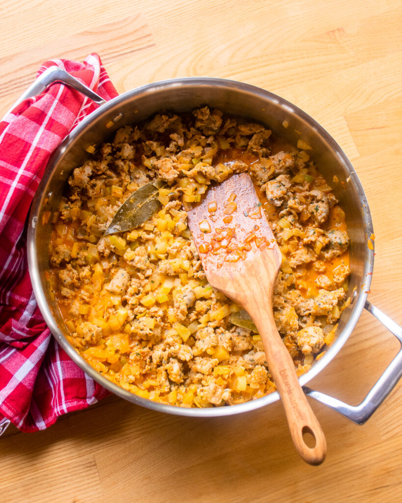 Looking down into a saute pan with a creamy meat sauce inside and a wooden spoon sticking out of it, with a red and white kitchen towel over one handle.