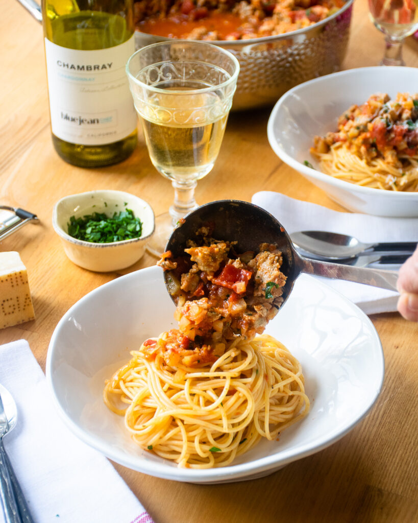 A ladle ladling sauce over a bowl of spaghetti with a glass and bottle of wine and a second place setting near by.