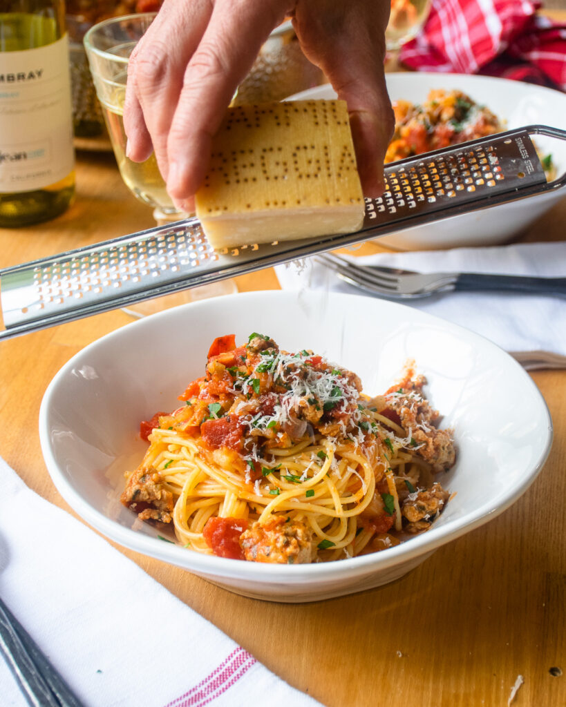 A hand grating Parmigiano-Reggiano over a bowl of pasta with chicken bolognese sauce.