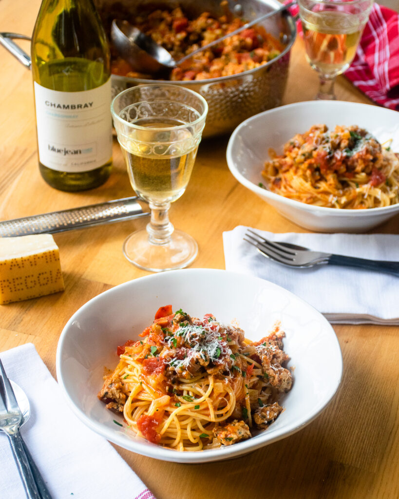 Two place settings with two white bowls of chicken bolognese over spaghetti, a bottle and glass of wine and a block of Parmesan cheese on the table.