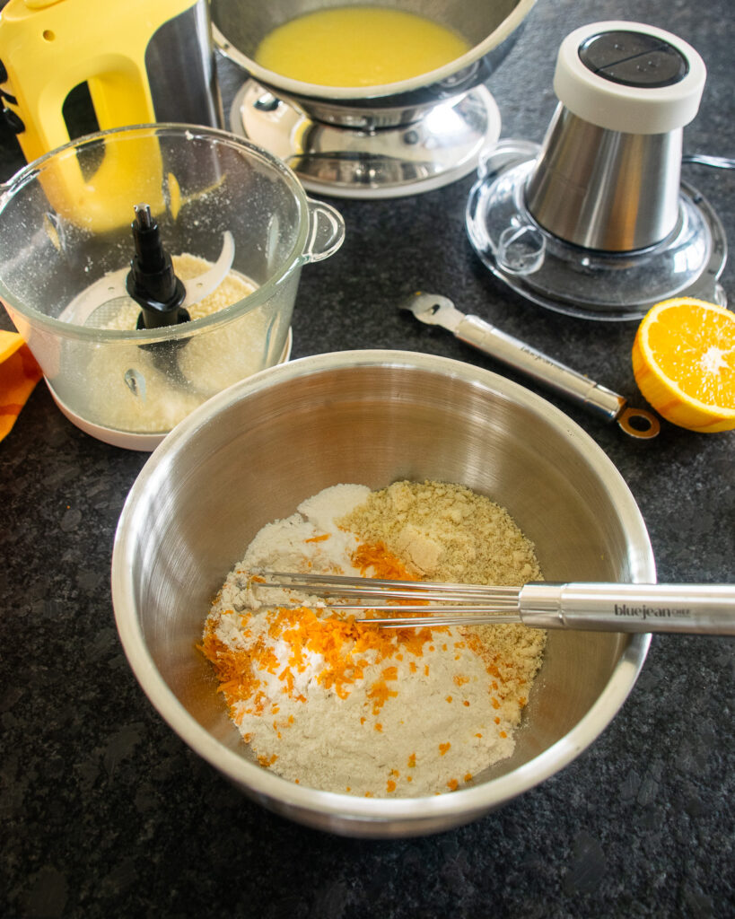 A whisk sticking out of a bowl with dry ingredients inside, a chopper in the background and other implements.