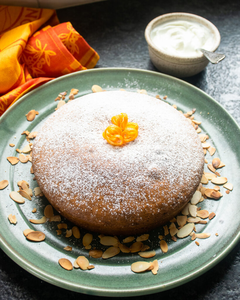 An orange almond olive oil cake on a green platter with toasted almonds around the side.