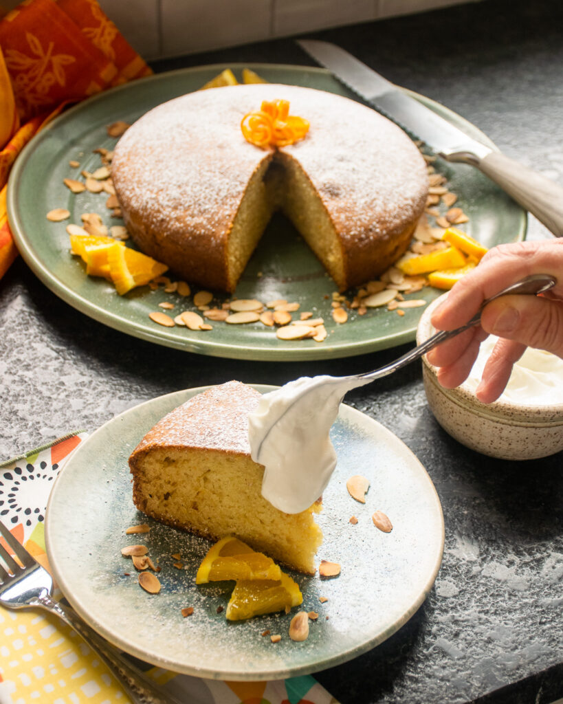 A hand dolloping yogurt onto a piece of orange almond olive oil cake.