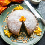 Orange Almond Olive Oil Cake with a piece cut out of it on a green platter with toasted almonds, orange segments and a bread knife.