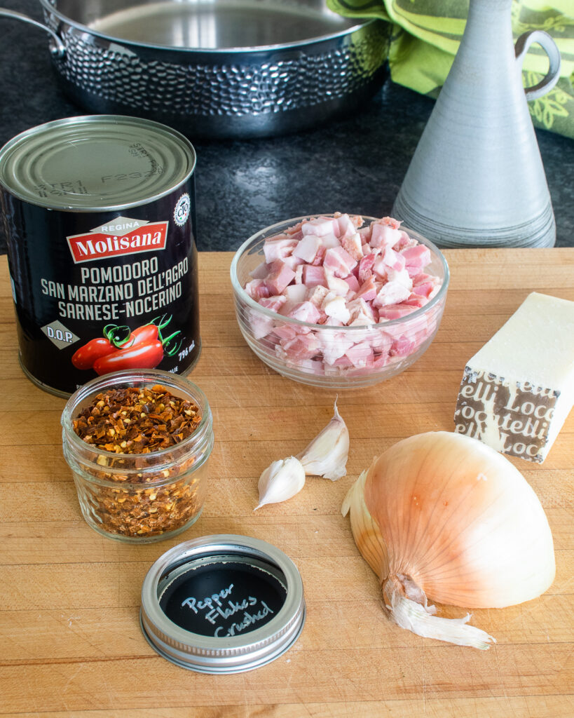 Ingredients on a cutting board - a can of tomatoes, chili flake, garlic, pancetta, half an onion and some pecorino cheese.