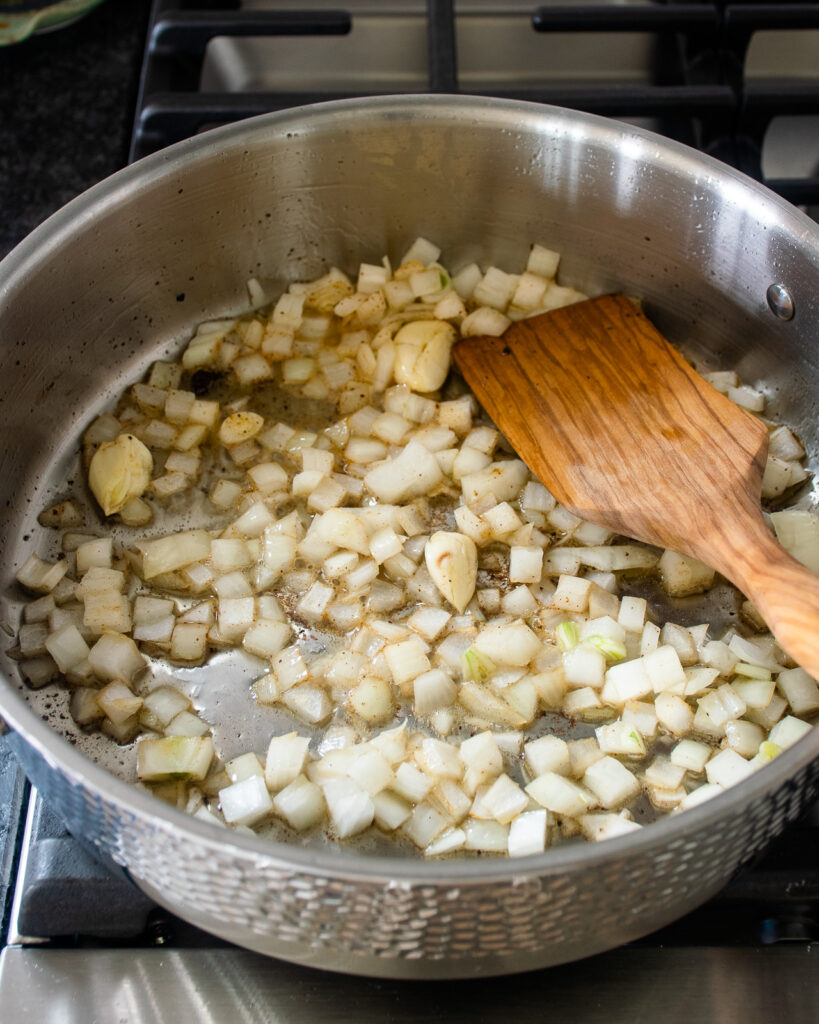 Onion and garlic cooking in a stainless steel saute pan with a wooden spoon sticking out.