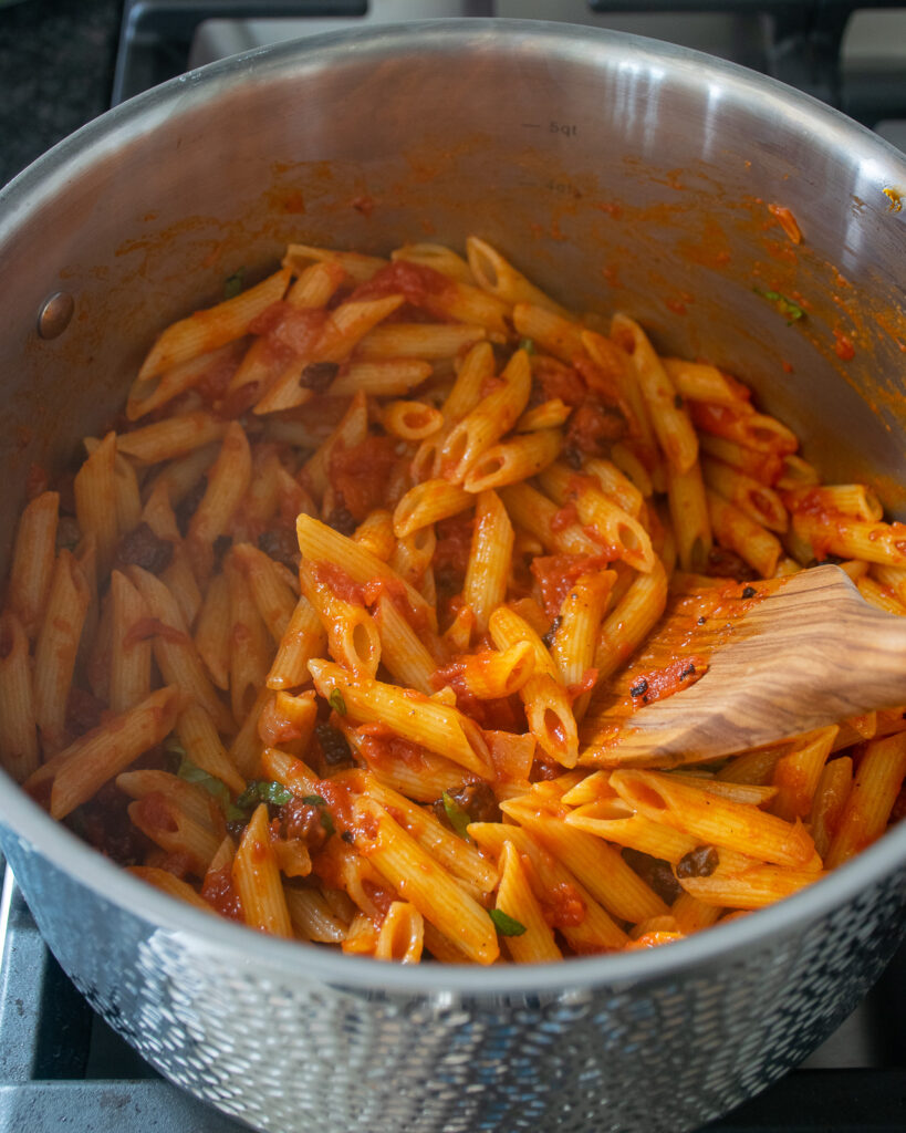 Penne amatriciana in a stainless steel stockpot with a wooden spoon.