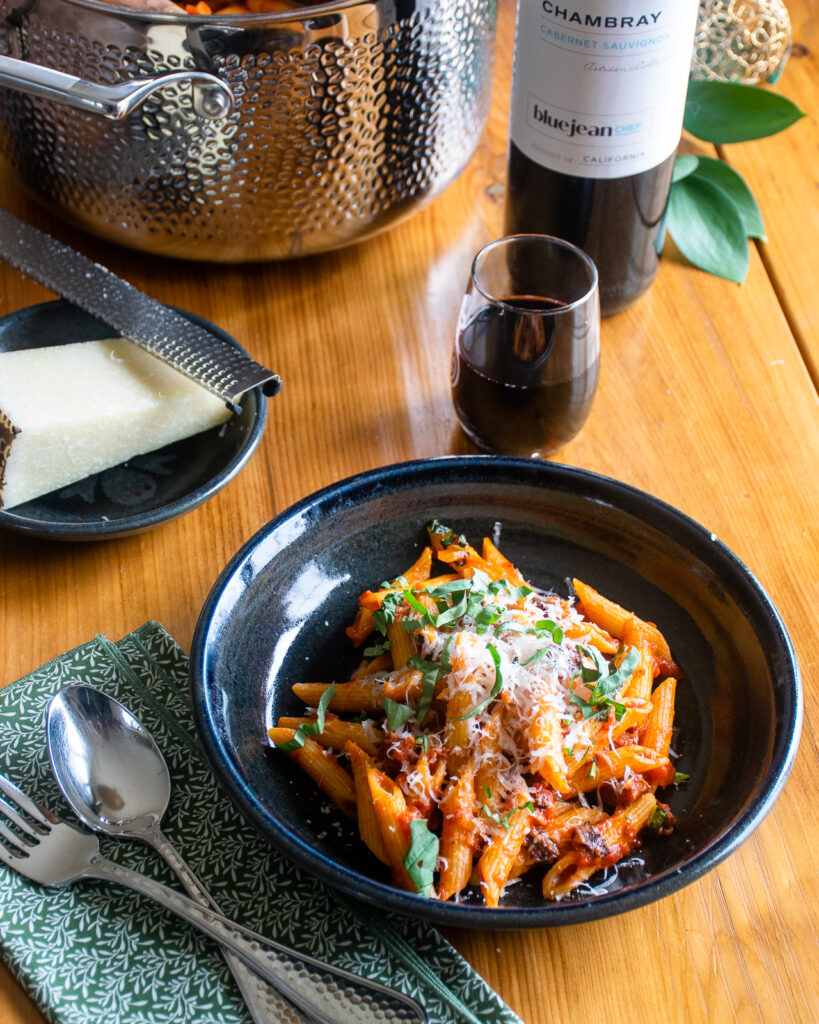 Pasta amatriciana in a black pottery bowl on a wooden table with a place setting and wine.