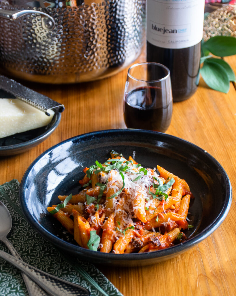 Pasta amatriciana in a black pottery bowl on a wooden table with a place setting and wine.