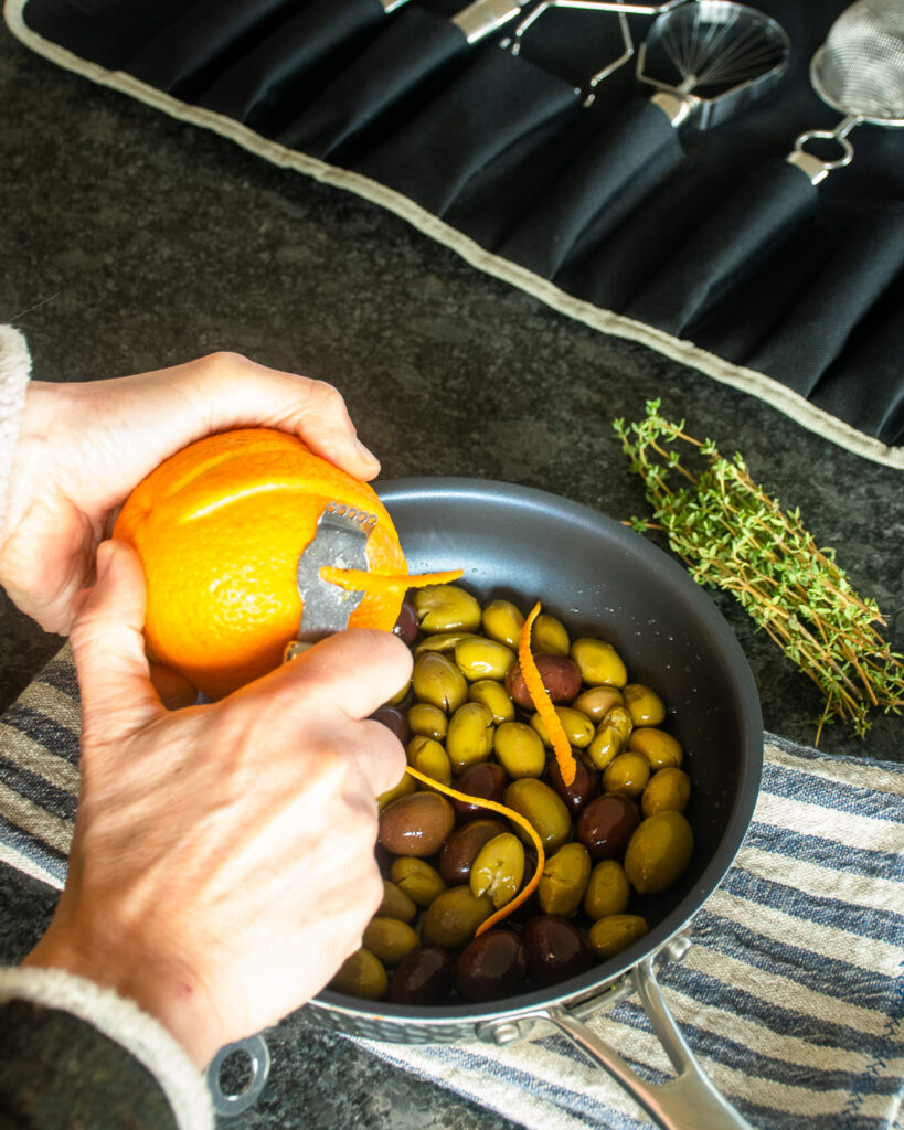 Hands using a channel knife to cut peel from an orange over a skillet with olives in it and thyme beside it. A gadget set in the background.