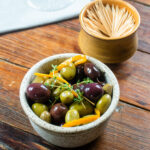 A ceramic dish of warm olives with a dish of toothpicks.