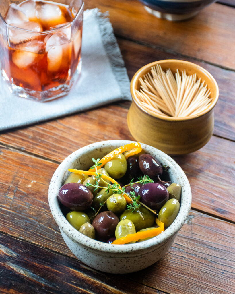 A ceramic dish of warm olives with a dish of toothpicks and a cocktail nearby.