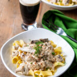 Beef stroganoff in a white bowl over egg noodles with a glass of Guinness in the background.