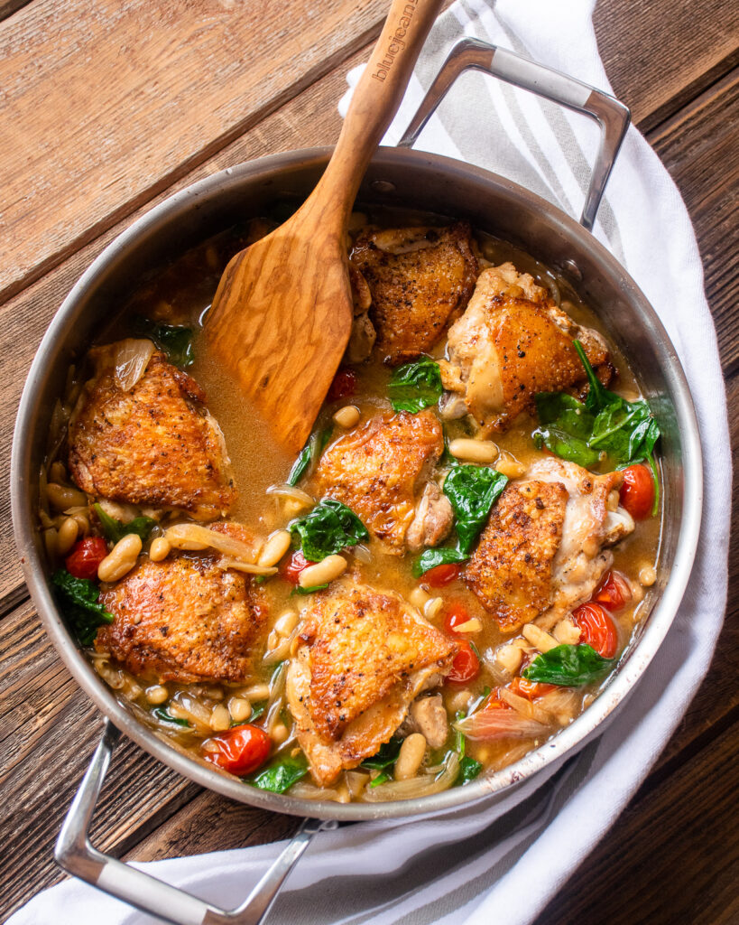 A stainless steel pan with crispy chicken thighs with white beans, tomatoes and spinach on a wooden table with a wooden spoon sticking out of it.