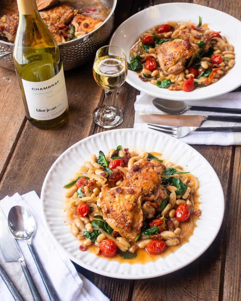 Two plates of chicken thighs with white beans and tomatoes on a wooden table with a saute pan, wine glass and bottle of wine.