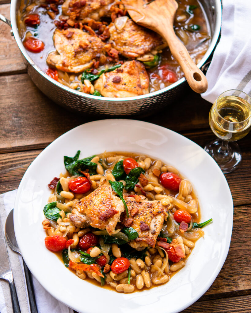 Looking down on a white plate and a saute pan both with chicken thighs with white beans and tomatoes in it.