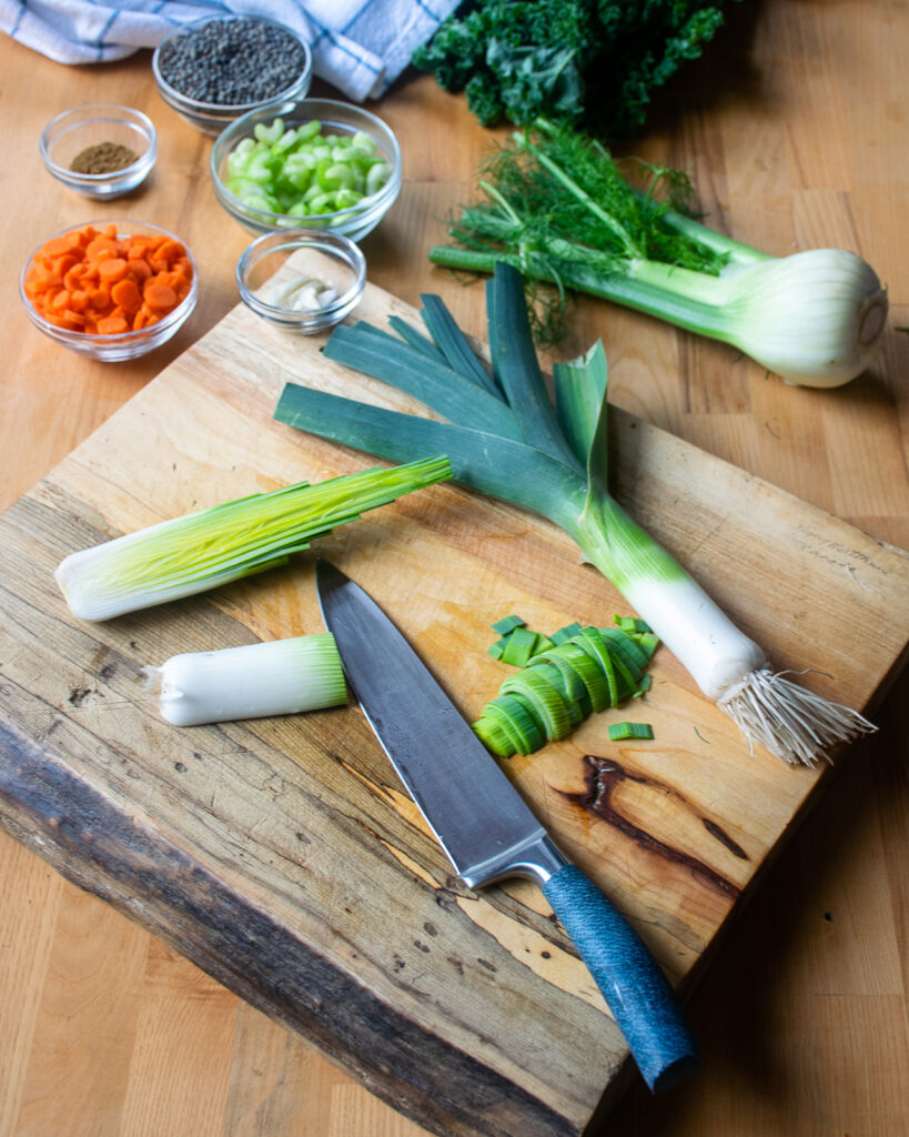 A leek being cut on a cutting board with other vegetables in various forms of prep around.