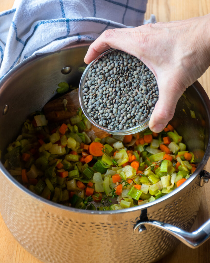 A hand holding a bowl of Puy lentils above a stockpot.