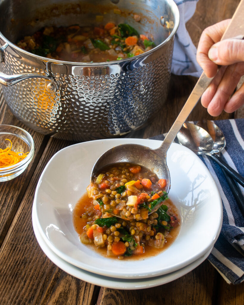 Ladling lentil soup into white bowls with a stockpot in the background.