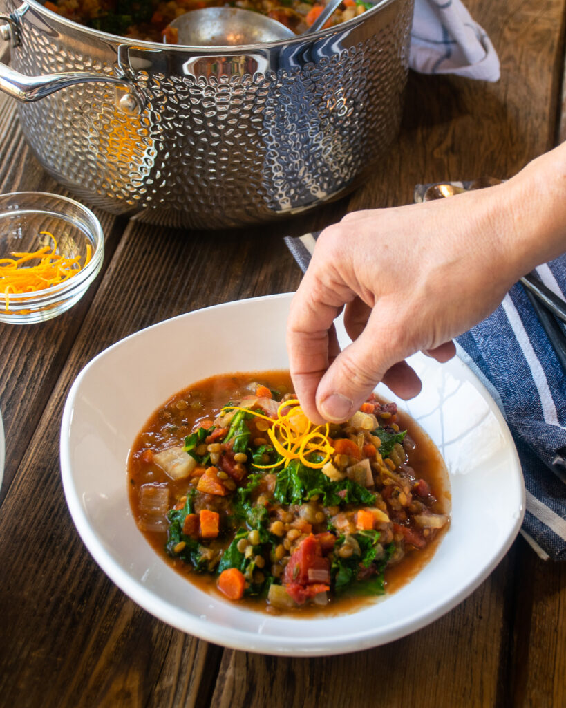 A hand garnishing a bowl of lentil soup with orange zest.