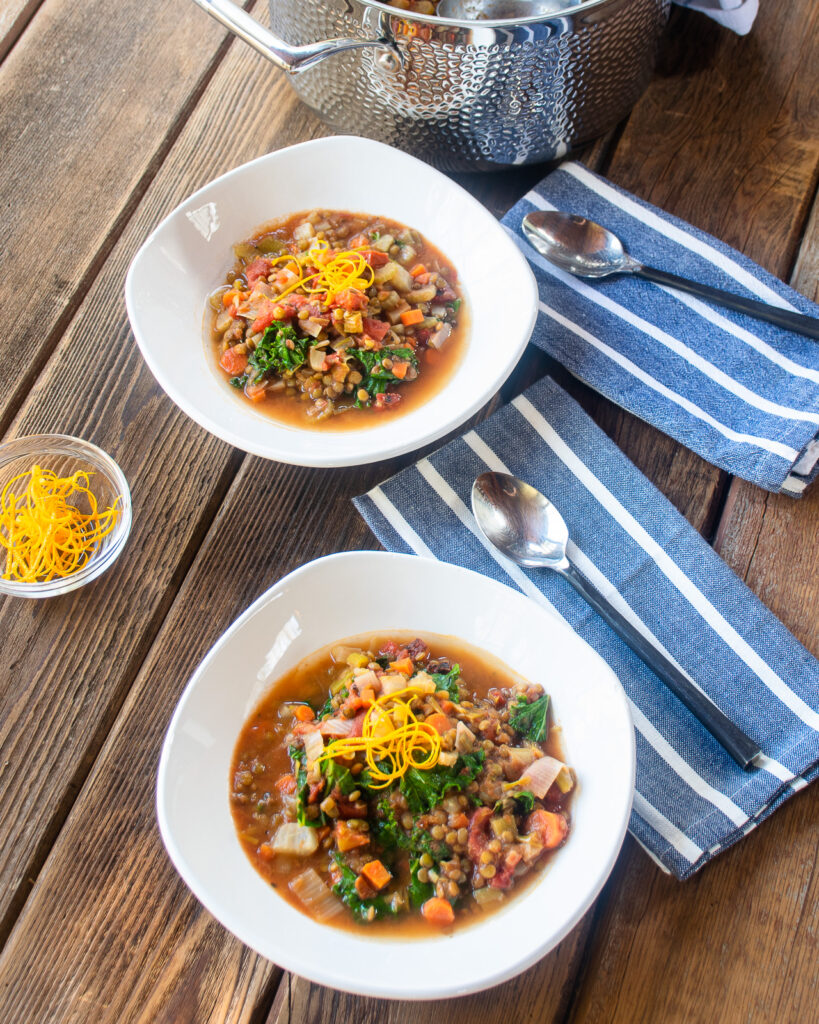 Two bowls of lentil soup on a wooden table with blue and white striped napkins and spoons. A stainless steel stockpot in the background.