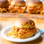 A spaghetti slider on a small plate in front of a wooden board with more sliders on it.