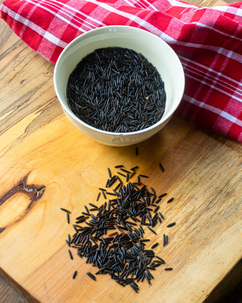 A bowl of raw wild rice on a cutting board with some rice on the cutting board and a red and white kitchen towel in the background.