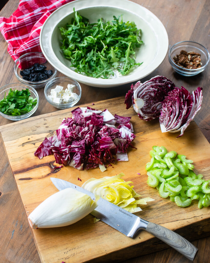 Various vegetables cut on a wooden cutting board with a bowl of arugula in the background.