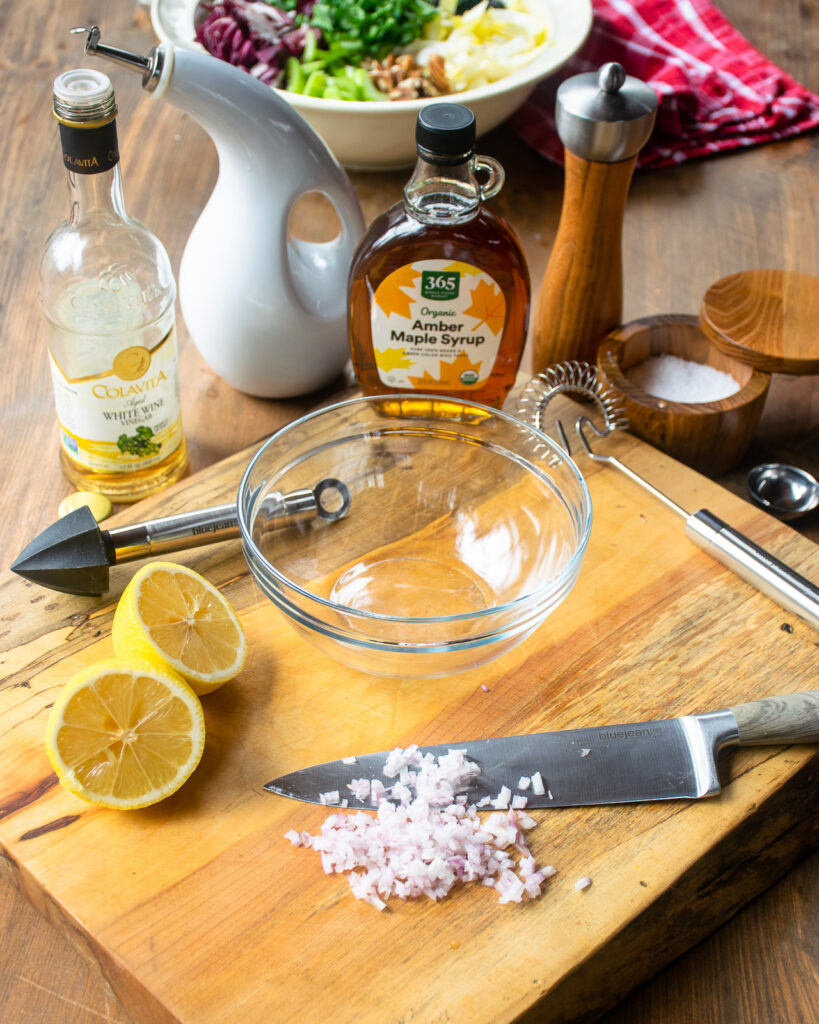 Ingredients for a salad dressing on a cutting board with a salad of various ingredients in the background.