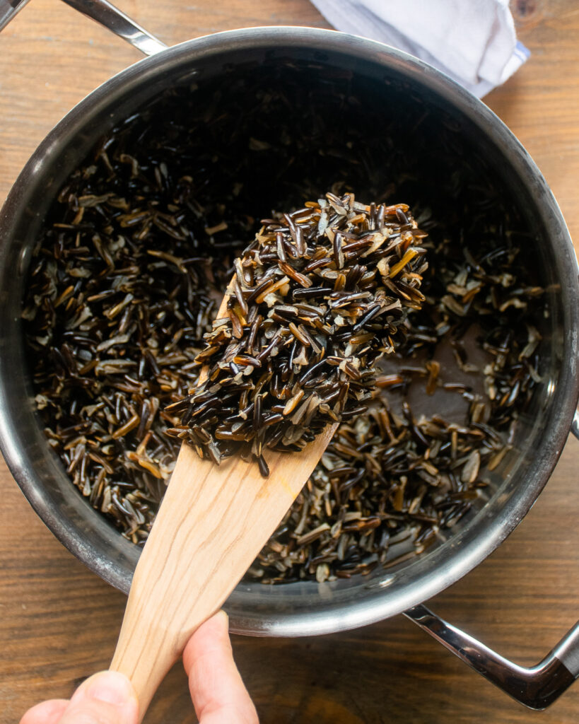 A wooden spoon holding cooked wild rice above a saucepan.