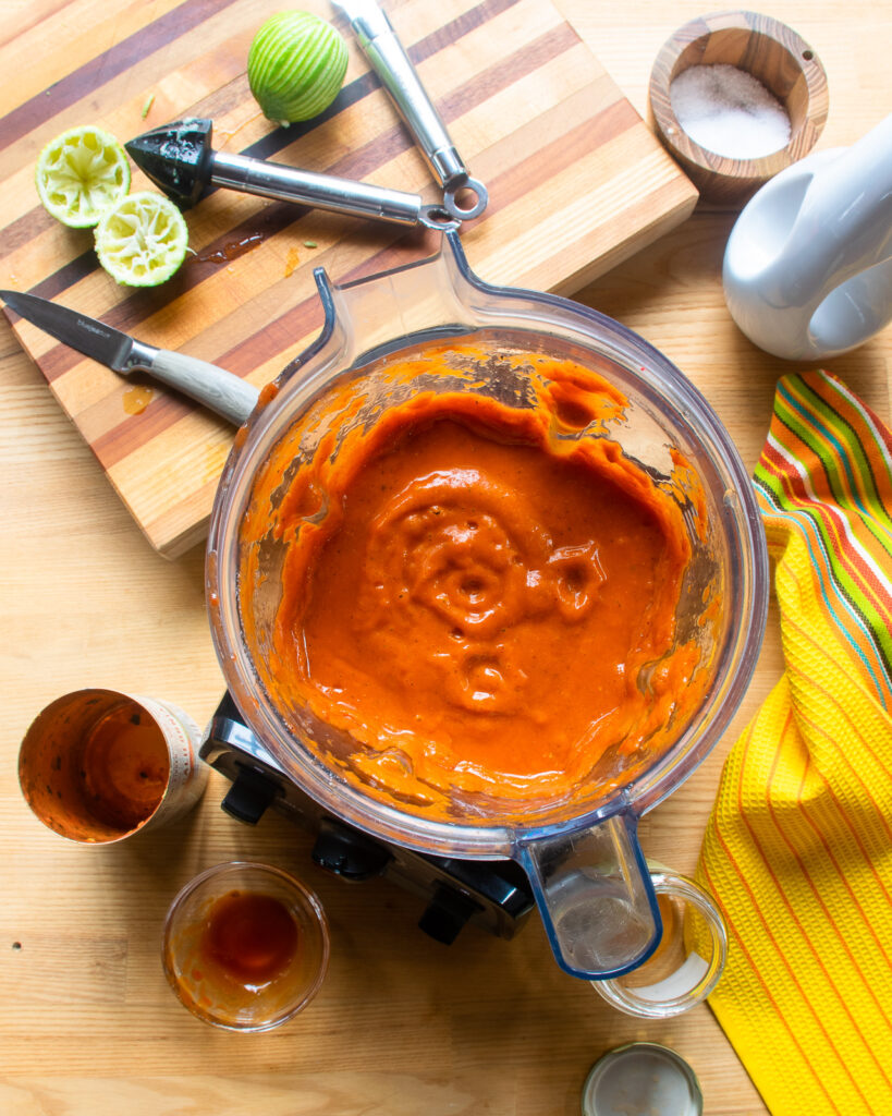 Looking down into a blender jar with enchilada sauce inside and various ingredients on the counter near by.