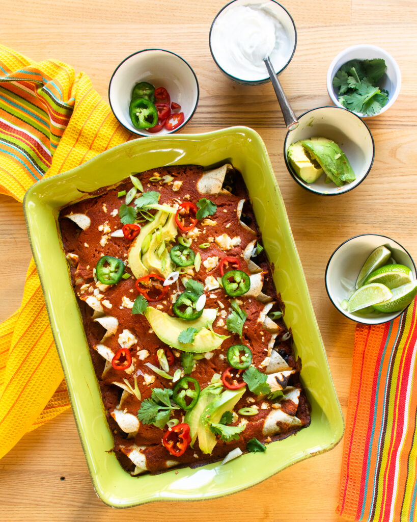 A green casserole of black bean enchiladas garnished with scallions, avocado, peppers.