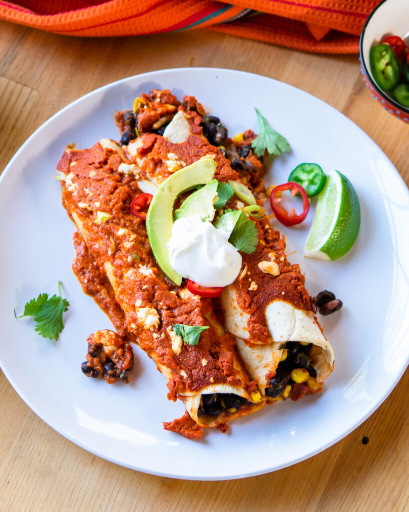 A white plate with black bean and corn enchiladas, avocado, sour cream, scallions and lime wedge.