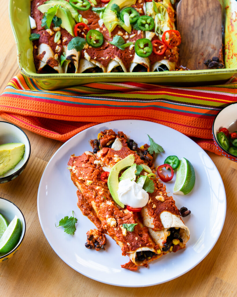 A white plate with black bean enchiladas in front of a green casserole dish.