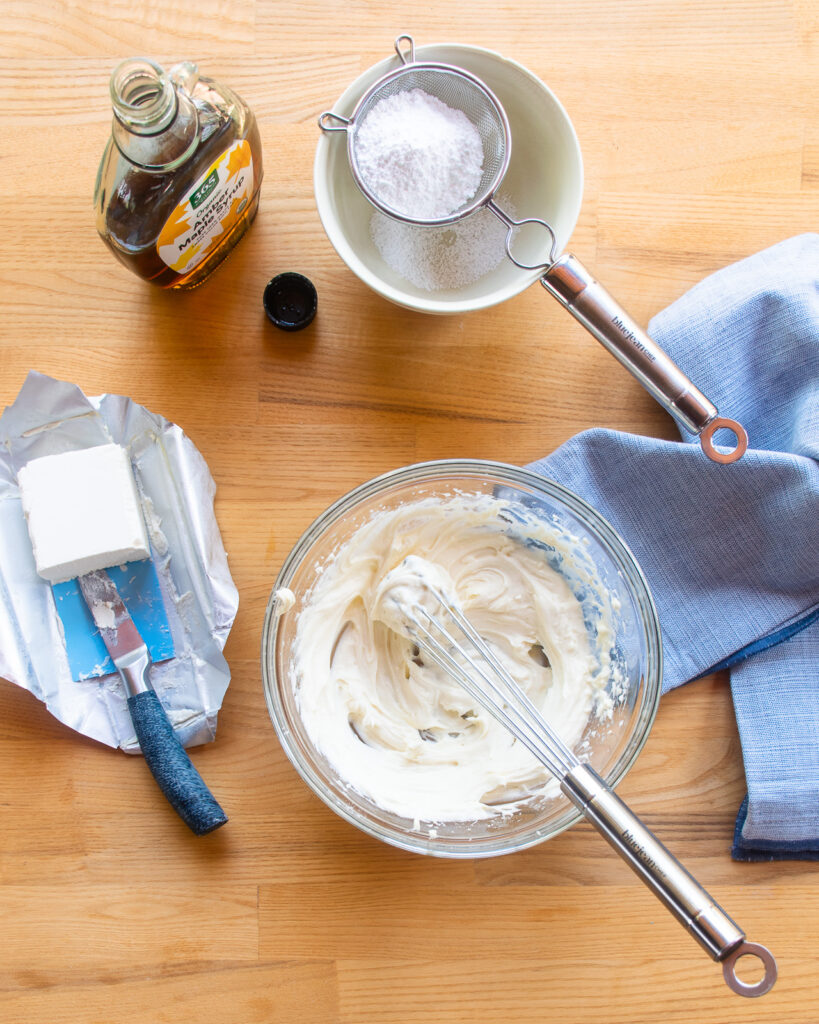 Looking down into a bowl of maple cream cheese with a whisk sticking out of the bowl and ingredients around.