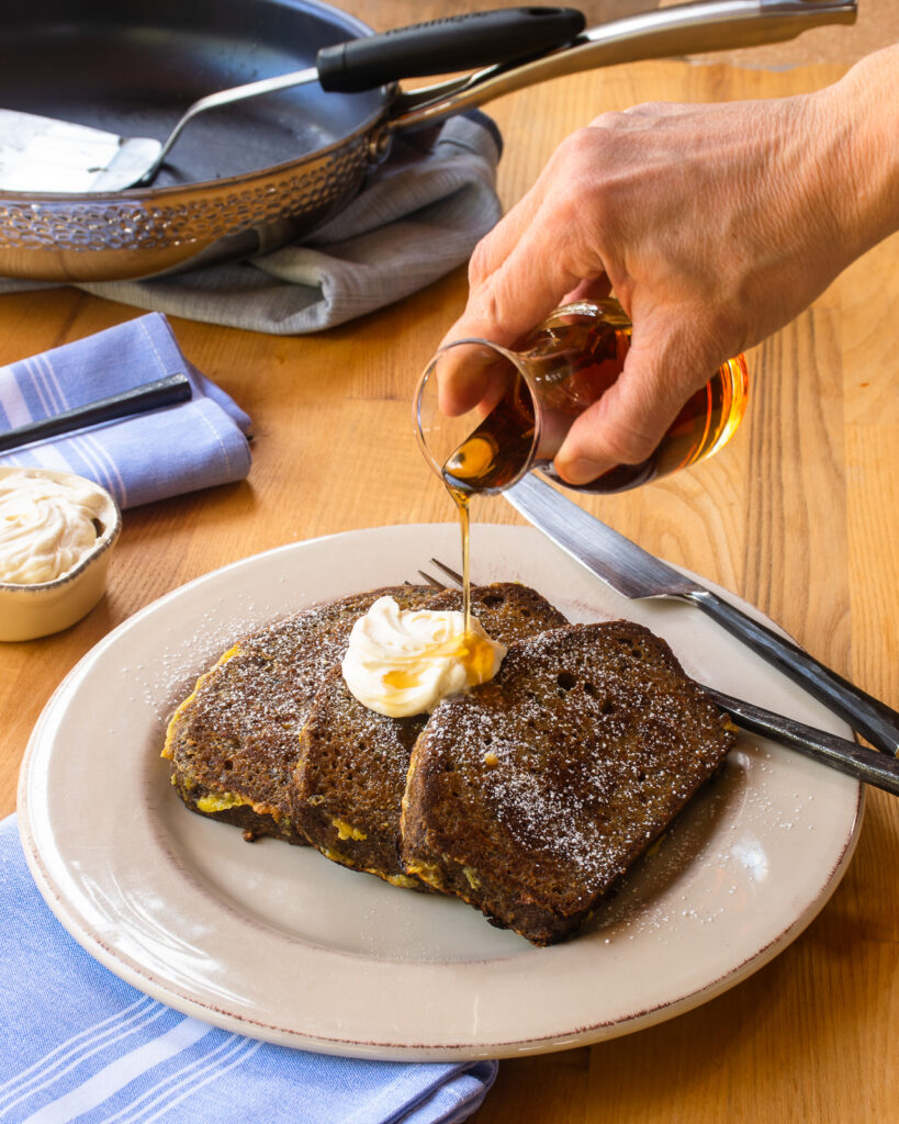 A hand pouring maple syrup over carrot cake french toast with a dollop of maple cream cheese on top.