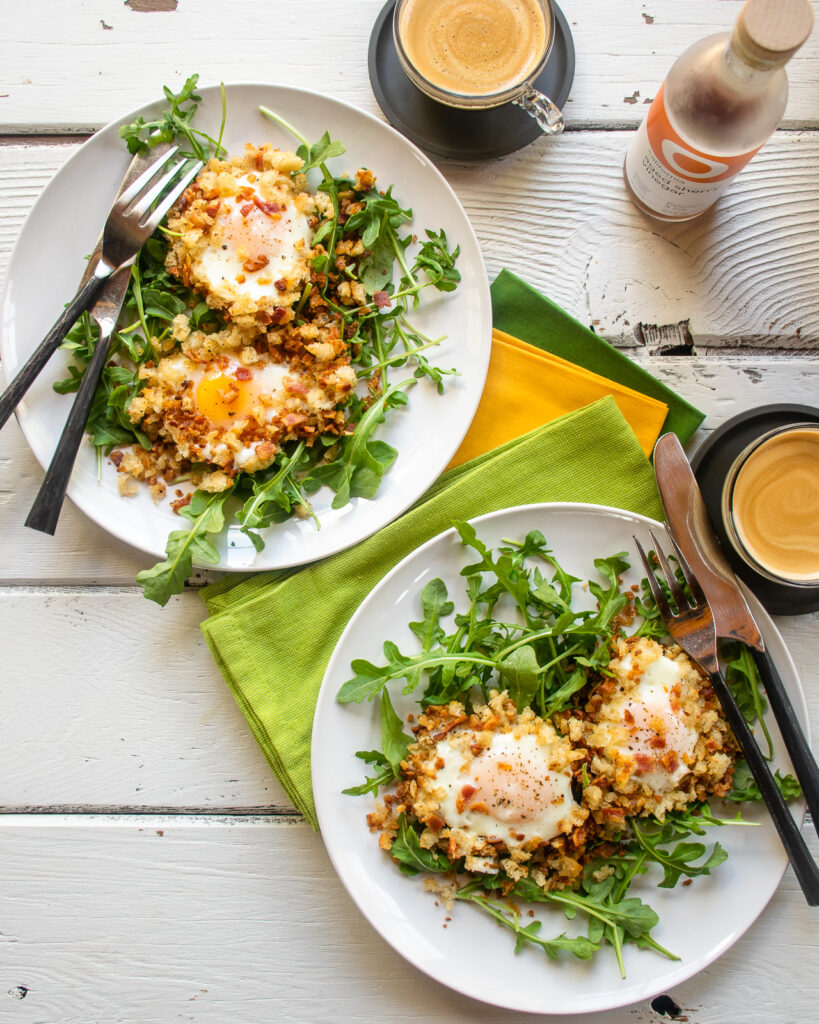 Two plates with crispy eggs in bacon breadcrumbs on a white table with coffees.